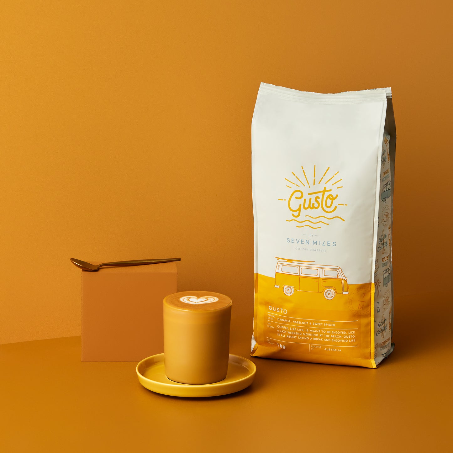 1x 1kg bag of Gusto Coffee by Seven Miles. Once glass coffee cup with a milky latte in the glass. A yellow box with a teaspoon on top.