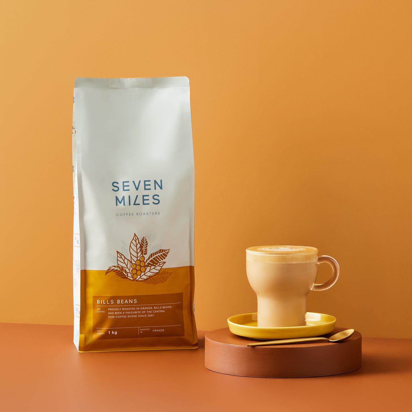 1x 1Kg bag of Mr Smooth Coffee by Seven Miles.  A brown circle coaster with a yellow saucer and glass coffee mug with gold teaspoon.