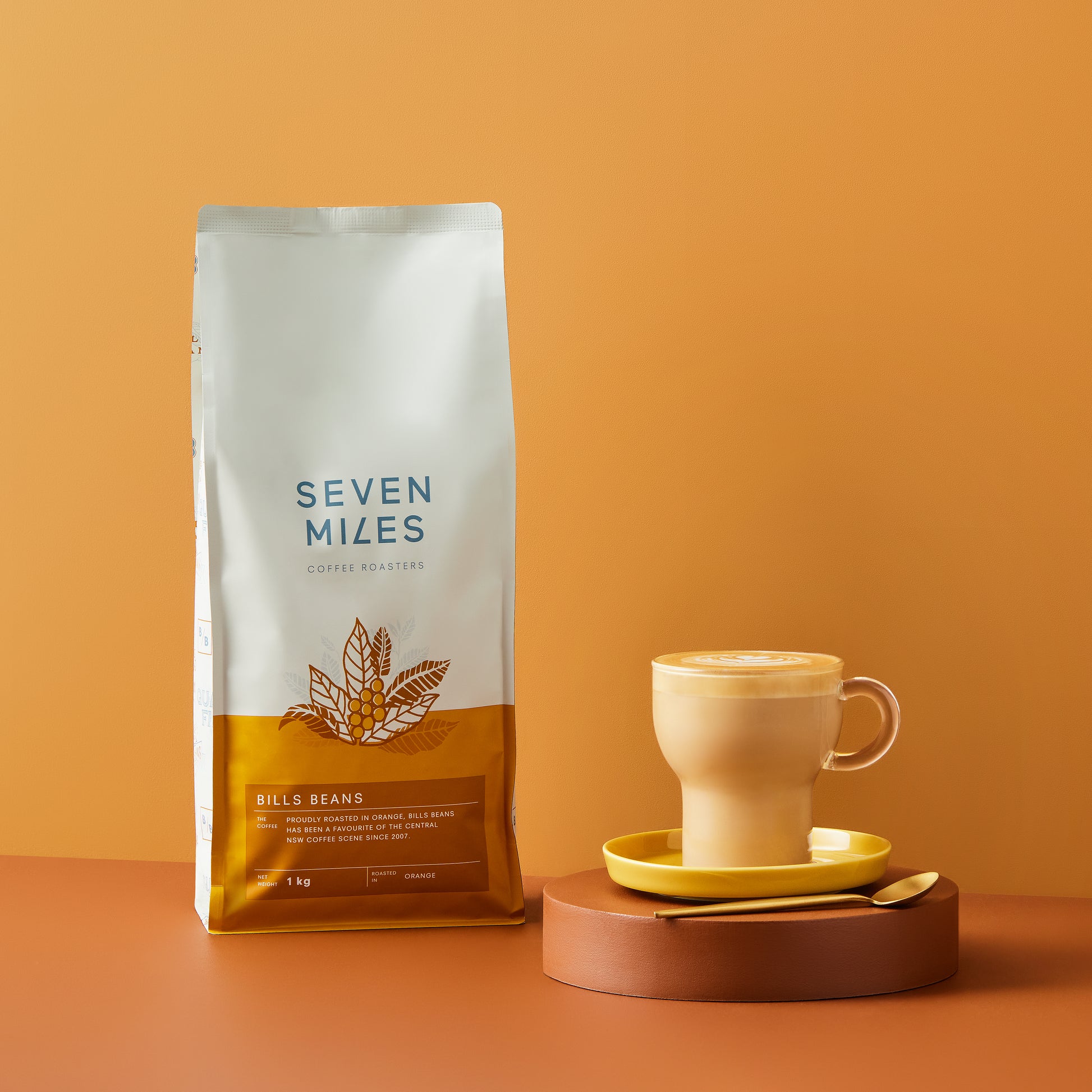 1x 1Kg bag of Mr Smooth Coffee by Seven Miles.  A brown circle coaster with a yellow saucer and glass coffee mug with gold teaspoon.