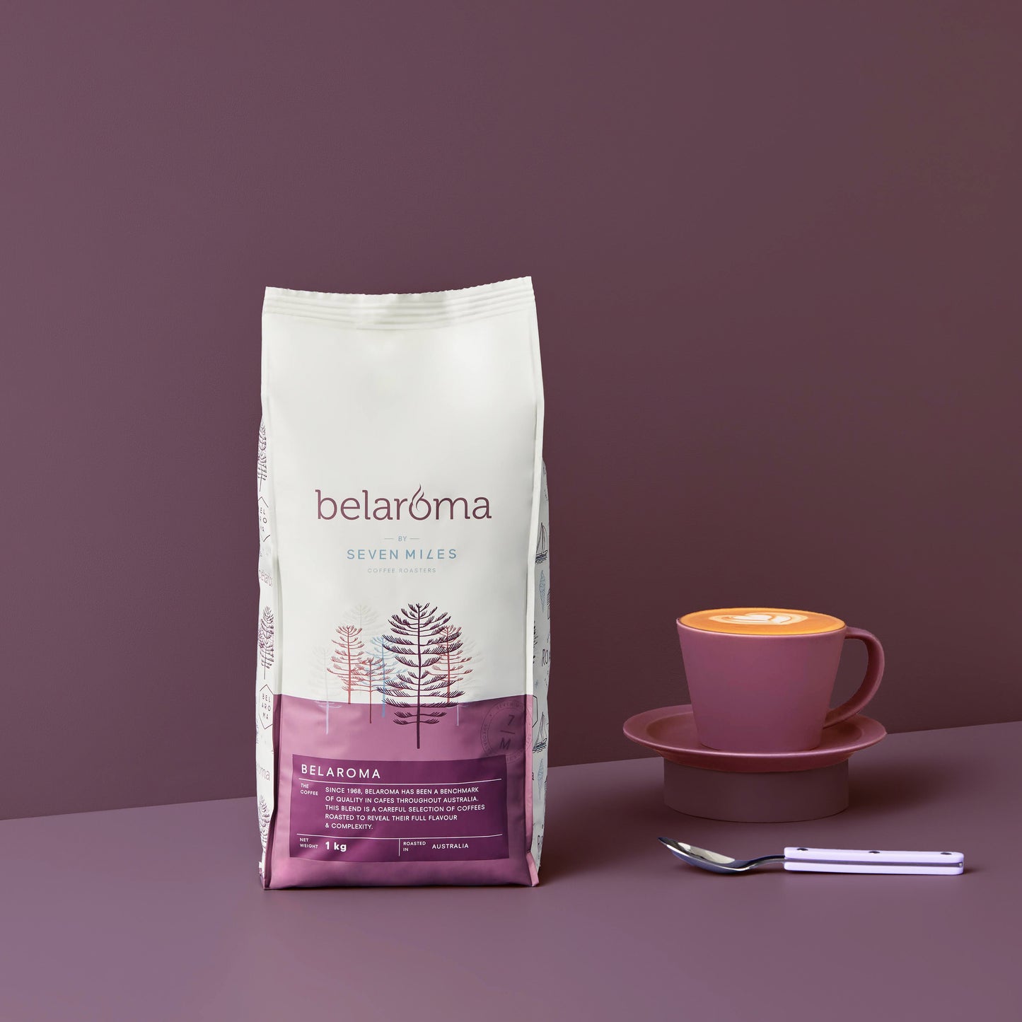 1kg bag of Belaroma Coffee by Seven Miles. Deep purple background with a purple mug and saucer and teaspoon.