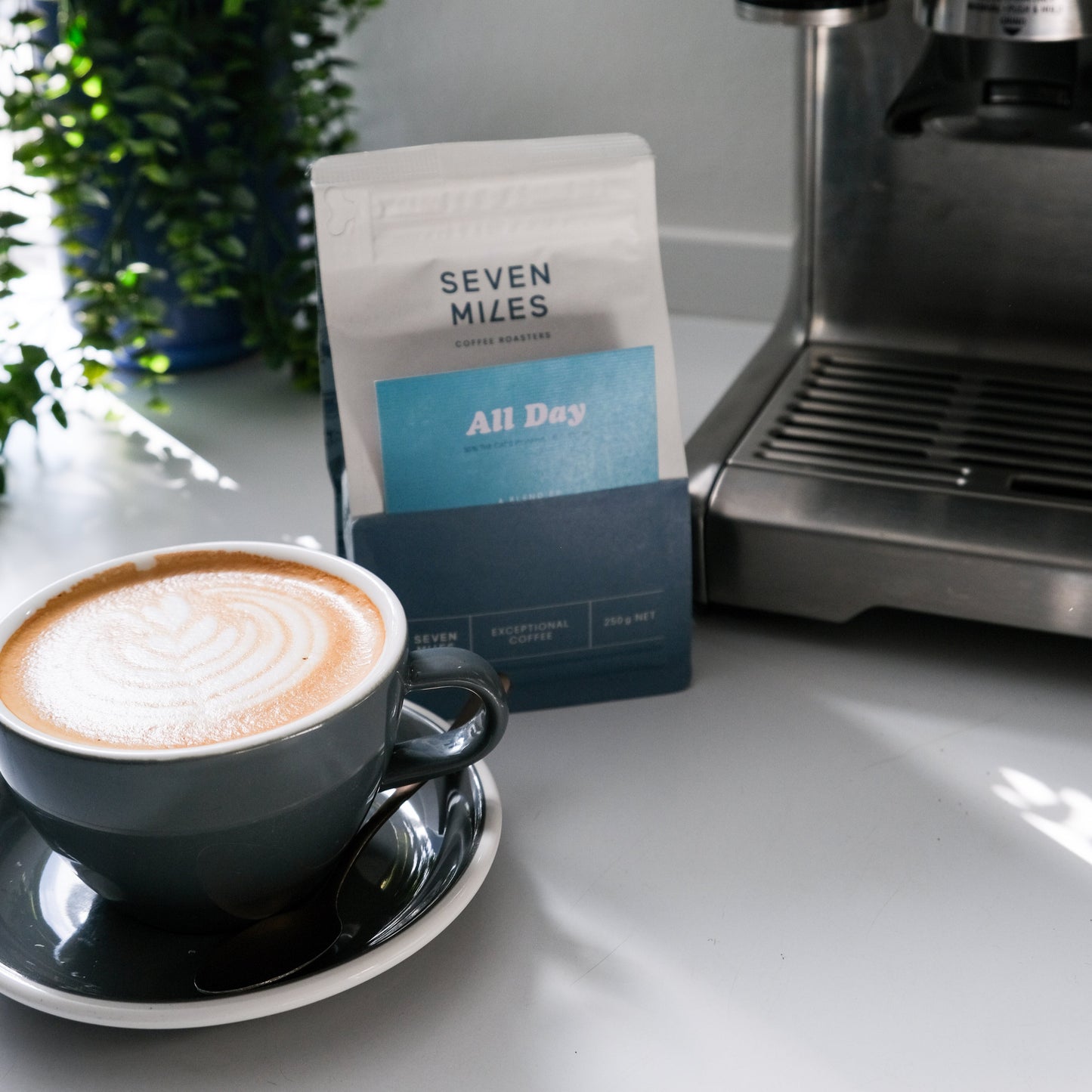 Cappuccino in a cup with a Seven Miles 250gr coffee bag of All Day on a kitchen counter next to an espresso machine.