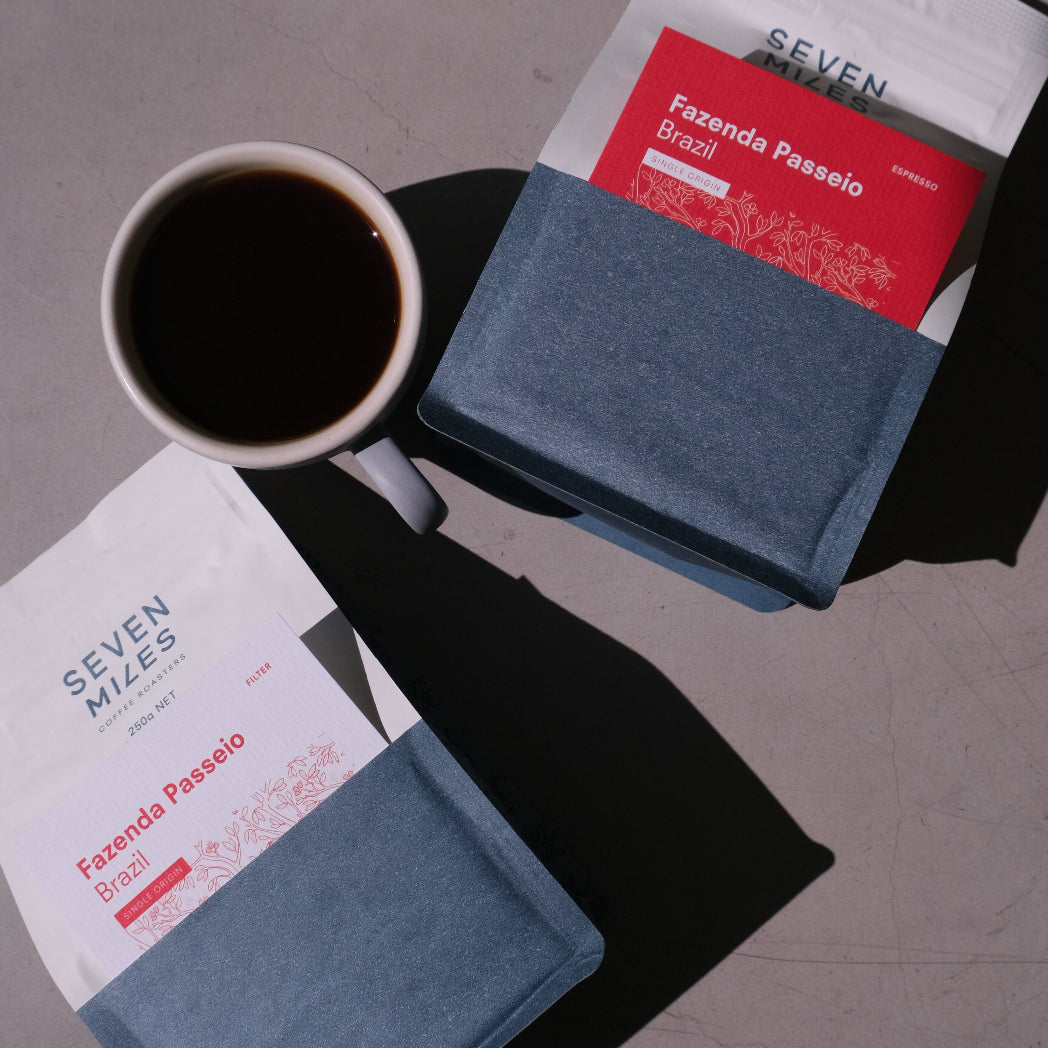 Two packages of Seven Miles Single Origin coffee and a cup of coffee on a gray surface.