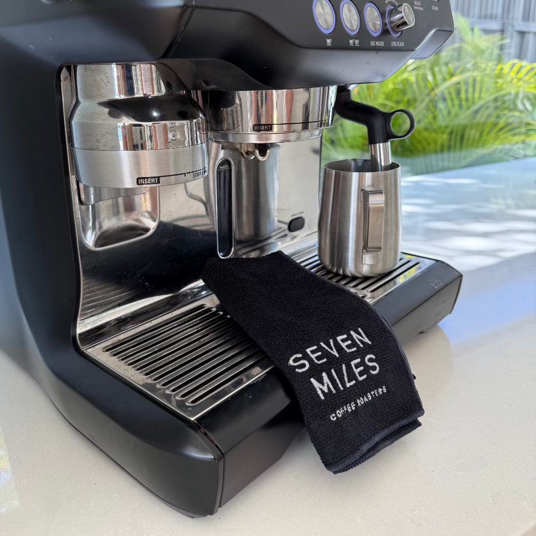 Black espresso machine on a kitchen counter with a hand holding a Seven Miles Coffee Cloth.