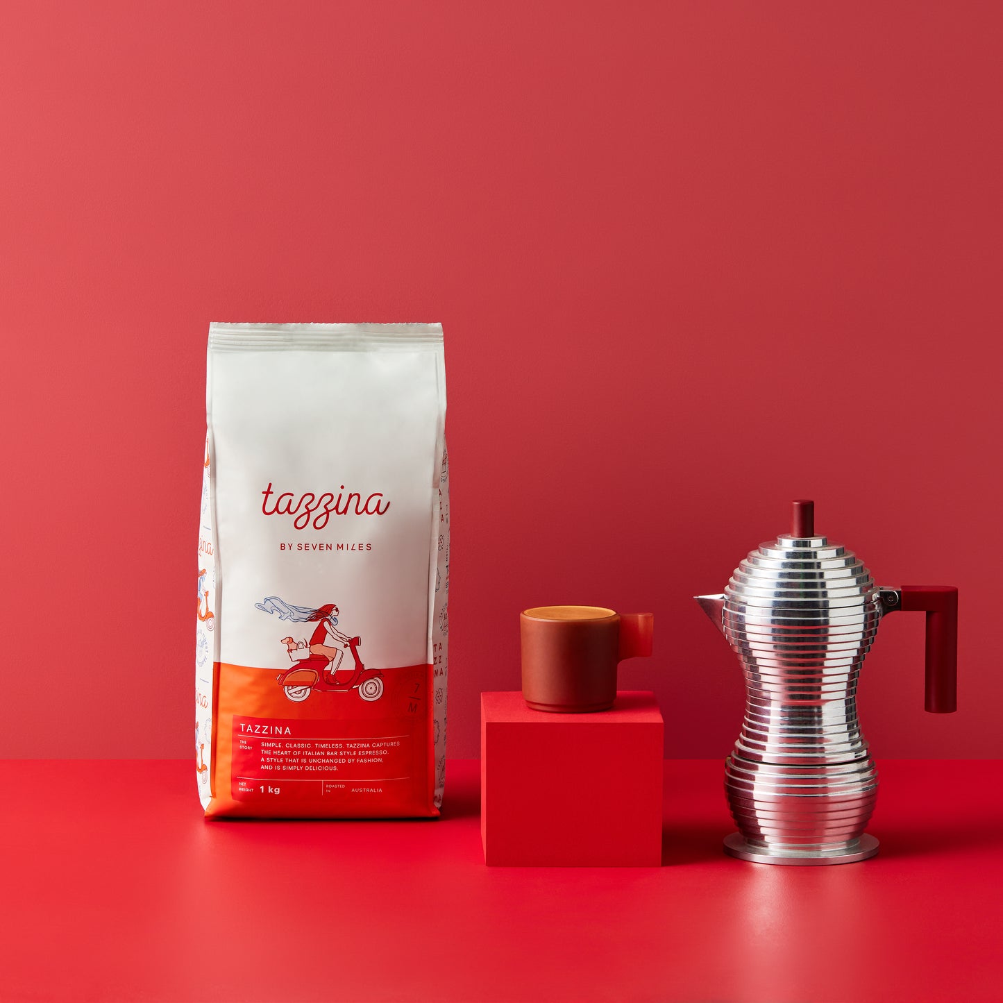 Tazzina Cafe 1kg coffee bag. One red glass coffee cup on a red box with a metal mokka pot. 