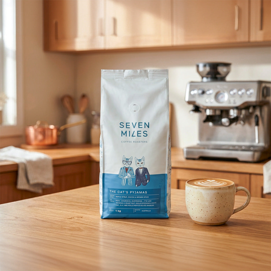 Seven Miles Coffee The Cat's Pyjamas 1kg bag featuring maple syrup, cocoa and brown spice notes, styled with latte art in home kitchen.