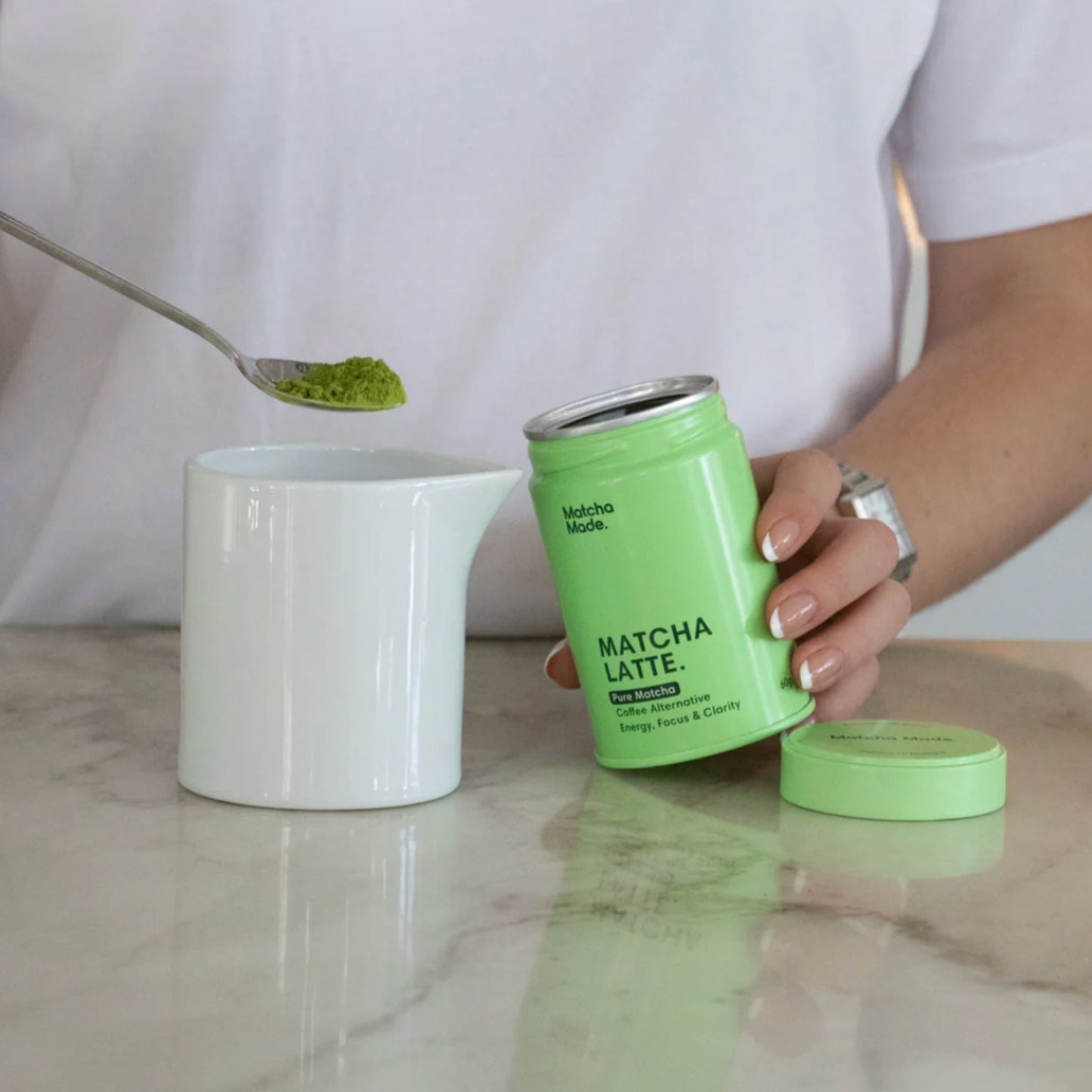 Person holding a green can labeled 'Matcha Latte' with a white mug and spoonful of matcha powder on a marble surface.