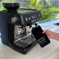 Black espresso machine on a kitchen counter with a hand holding a Seven Miles Coffee Cloth. 