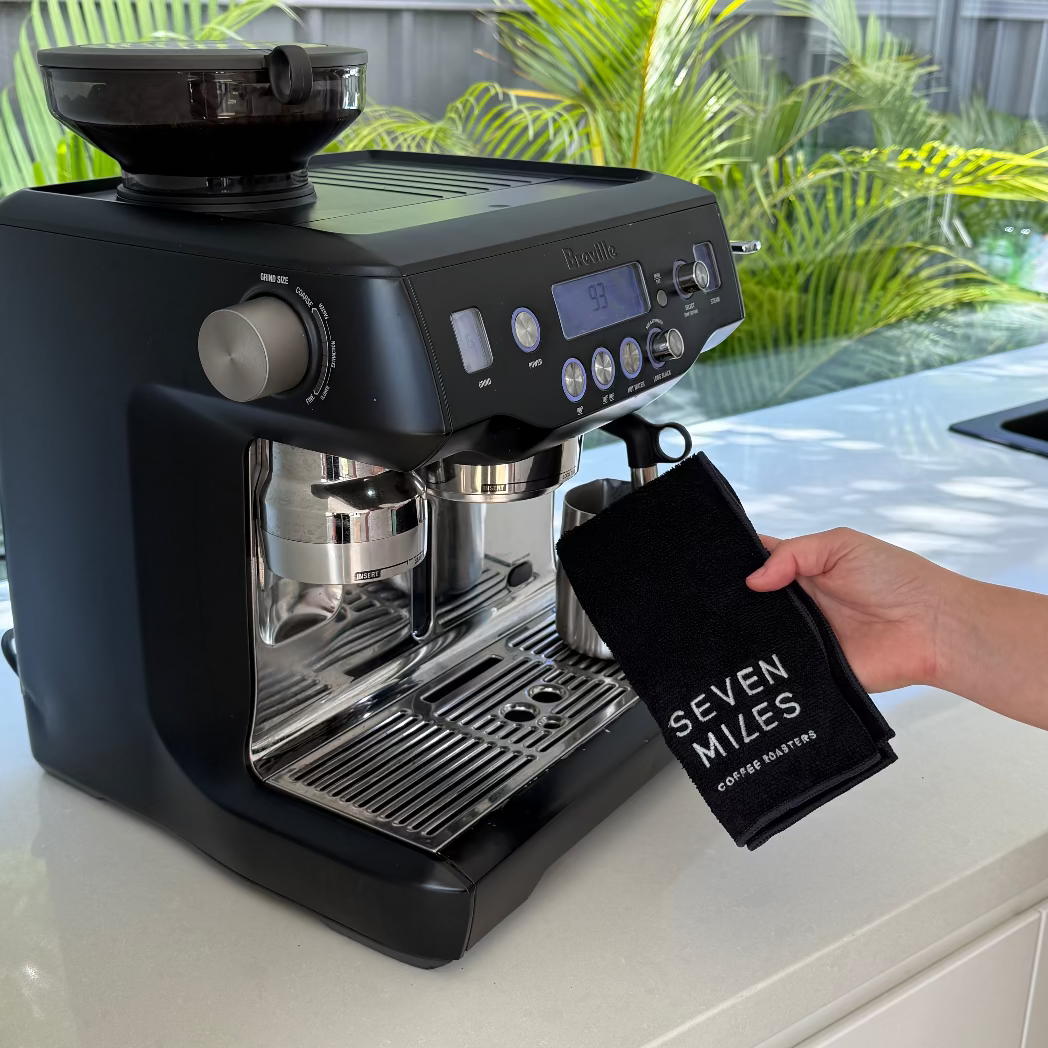 Black espresso machine on a kitchen counter with a hand holding a Seven Miles Coffee Cloth. 