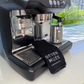 Black espresso machine on a kitchen counter with a hand holding a Seven Miles Coffee Cloth. 